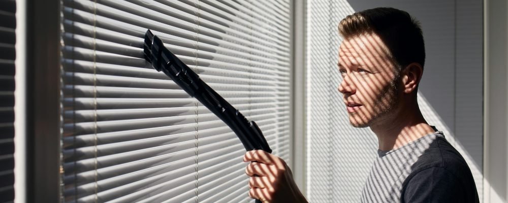Man cleaning dust from window blind by vacuum cleaner